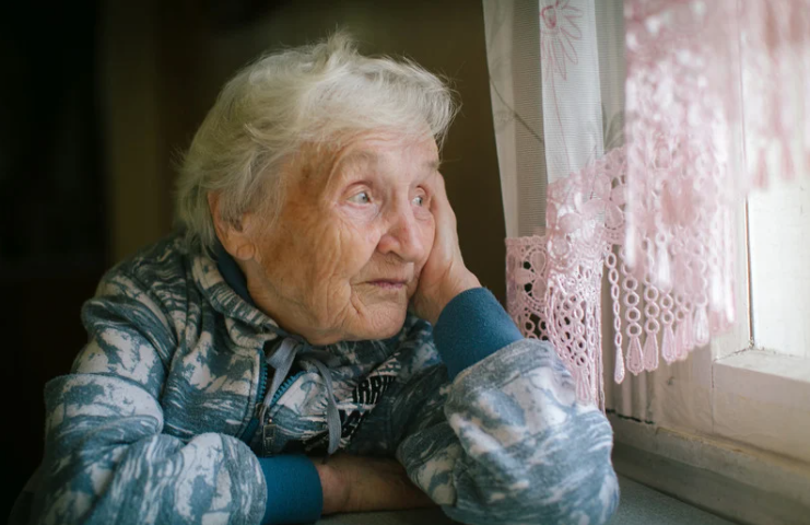 Elderly woman sitting alone looking out a window, illustrating the isolation that puts seniors at higher risk of phone and online scams